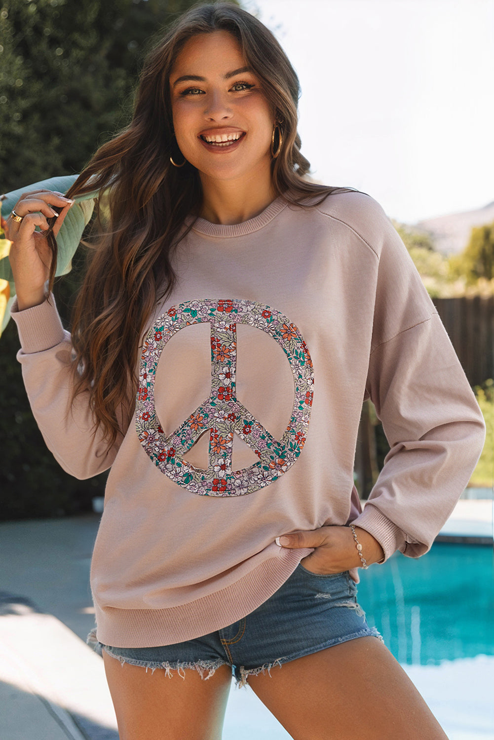 Woman wearing a beige sweatshirt with a floral peace symbol design, standing by a pool.